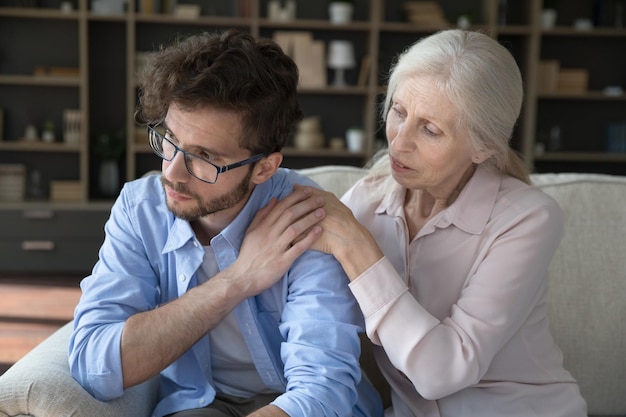 Mère âgée préoccupée parlant à son fils adulte frustré et déprimé, réconfortant, soutenant la famille, touchant les épaules, aidant à faire face aux problèmes, perte, assis près du canapé à la maison.