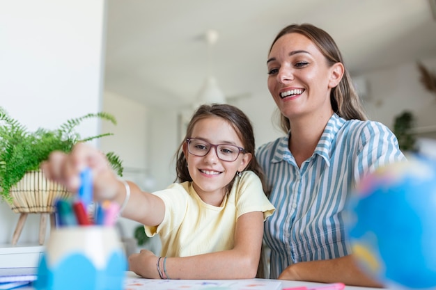 Mère aidant sa fille tout en étudiant à la maison. Jolie femme souriante aidant une fille adorable à faire ses devoirs à la maison