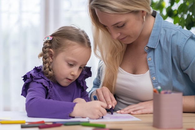 La mère aide sa fille à faire ses devoirs ou ses devoirs scolaires ensemble assis au bureau dessinant une image