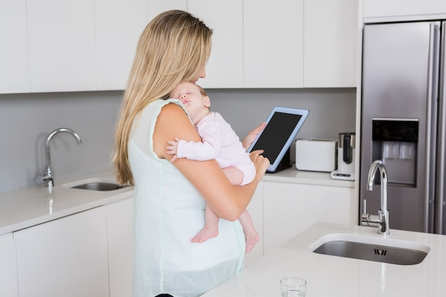 Mère à l'aide de tablette numérique tout en portant son bébé dans la cuisine