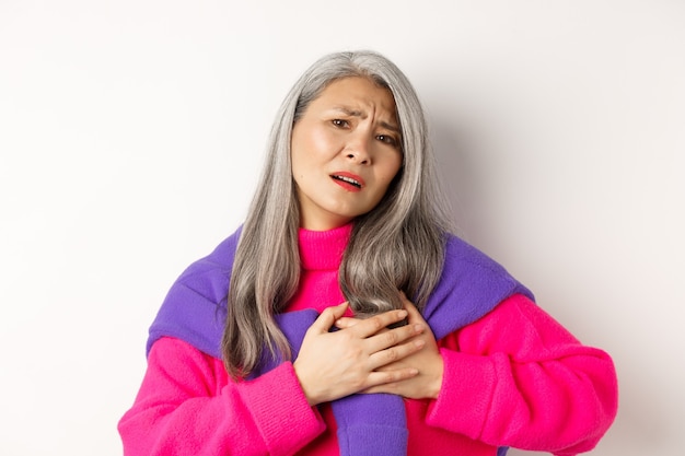 Une mère asiatique inquiète tenant la main sur le cœur et grimaçant, l'air anxieuse et ayant mal à la poitrine, se sentant blessée, debout sur fond blanc