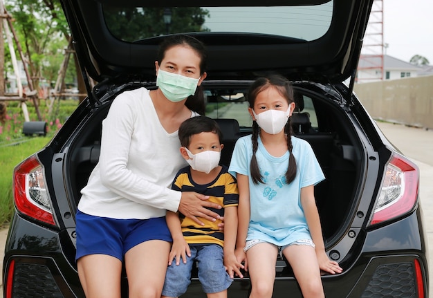 Une mère asiatique et un petit garçon et une fillette portent un masque d'hygiène assis sur une voiture à hayon en regardant à travers la caméra pendant l'épidémie de coronavirus (covid-19)