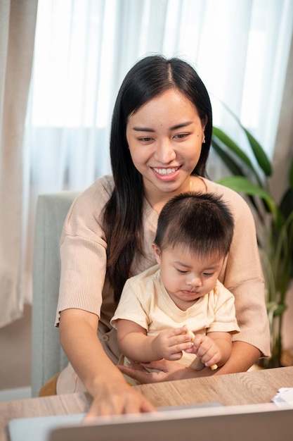 Une mère asiatique positive s'occupe de son petit garçon tout en travaillant sur son ordinateur portable