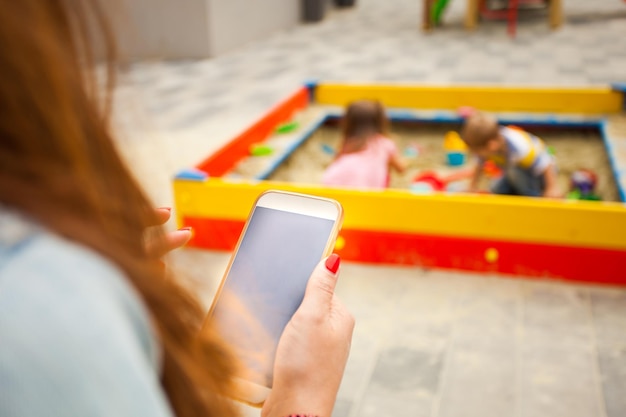 Mère assise et regardant l'écran du smartphone. Discuter ou bloguer et s'occuper des enfants. Les enfants jouent au bac à sable sur le fond