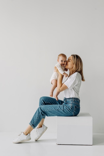 Mère assise et tenant bébé dans les bras souriant et jouant sur fond de mur blanc