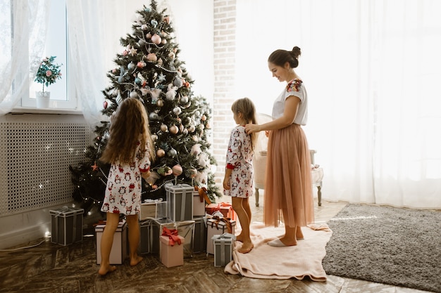Une mère attentionnée tresse la tresse de sa petite fille tandis que la deuxième fille décore un arbre du Nouvel An dans la pièce lumineuse et confortable.