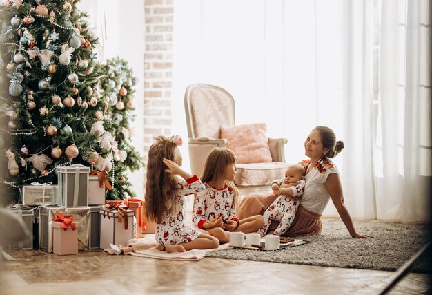 Une mère avec un bébé sur les mains est assise sur le tapis avec ses deux filles en pyjama en train de manger des biscuits au cacao avec des guimauves près de l'arbre du Nouvel An dans la chambre confortable .