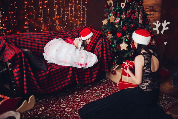 Mère en belle robe noire et chapeau de père Noël avec présent contre enfant endormi sur le canapé.