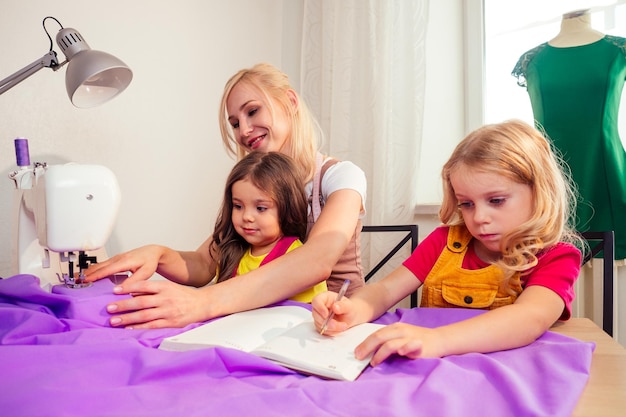 Une mère blonde souriante et multitâche apprend à ses petites sœurs à coudre seule sur une machine à coudre