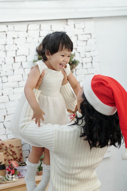 Mère en bonnet de noel soulevant une petite fille riante en robe blanche