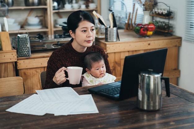 mère de carrière asiatique travaillant à domicile boit du café tout en regardant l'écran de l'ordinateur portable avec sa petite fille assise dans les bras à table dans la salle à manger.