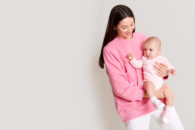 Mère caucasienne aux cheveux noirs tenant bébé dans les mains et regardant sa fille, belles robes féminines rose sweat-shirt avec bébé isolé sur un mur blanc avec espace de copie.