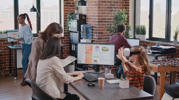 Mère célibataire avec enfant au bureau, fille s'amusant avec un casque VR pendant que maman travaille avec un collègue. Parler de graphiques commerciaux sur ordinateur et presse-papiers. Maman célibataire faisant un travail et m