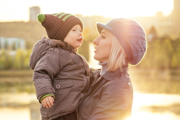 Mère dans une veste chaude et un bonnet tricoté étreignant son petit garçon marchant dans le parc d'automne femme blonde élégante avec un enfant sur fond de lac