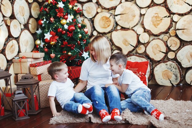 Mère et deux garçons en chapeaux de Noël sont assis près de l'arbre du Nouvel An à l'arrière-plan des coupes d'arbres.