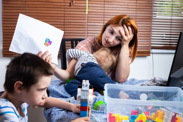 Mère avec deux petits garçons à genoux essaie de rire à la maison. jeune femme s'occupe des enfants et travaille sur un ordinateur.