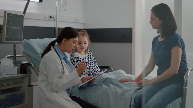 Mère discutant avec une petite fille malade pendant qu'une infirmière afro-américaine arrange le lit pendant l'examen médical dans la salle d'hôpital. Médecin médecin expliquant les symptômes de surveillance du traitement de la maladie