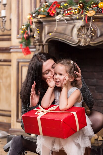 Mère embrassant sa petite fille mignonne avec un cadeau de Noël près d'une cheminée avec des décorations