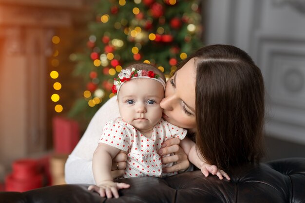 Une mère embrasse un enfant à Noël dans une pièce avec des décorations, un sapin de Noël et des lumières