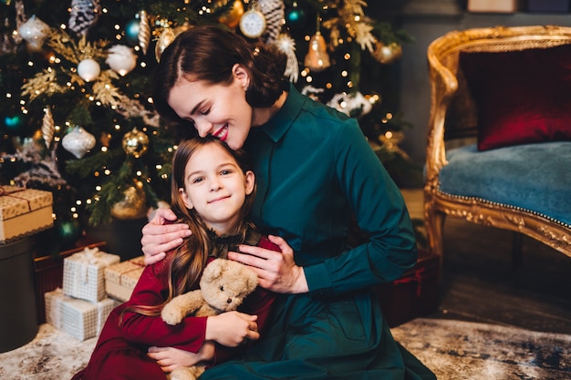 Mère embrasse sa petite fille comme assis ensemble près d'un arbre de Noël décoré