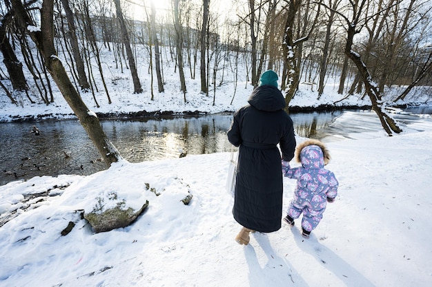 Mère et enfant marchant sur une journée d'hiver glaciale ensoleillée dans le parc près de la rivière avec des canards et des oiseaux