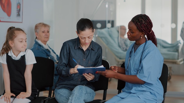 Mère avec enfant signant des papiers de contrôle dans la salle d'attente, parlant à l'infirmière du diagnostic et du traitement. Femme recevant des conseils médicaux d'un assistant de santé dans le hall de l'établissement.