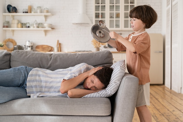 Une mère fatiguée et épuisée couvre les oreilles allongée sur un canapé avec un fils bruyant qui s'ennuie fait du bruit des ustensiles de cuisine