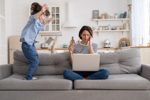 Mère fatiguée s'asseoir sur le canapé, travailler sur un ordinateur portable à la maison, petit enfant hyperactif sautant en attirant l'attention.