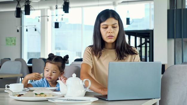 La mère d'une femme d'affaires asiatique travaille assise à table avec du thé et un ordinateur portable gris et l'enfant mange une tranche de pizza au tout-petit