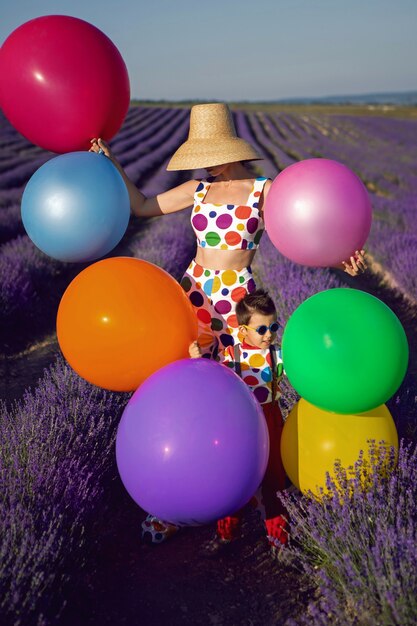 Mère de femme extravagante dans un costume blanc à pois multicolores et un chapeau se dresse dans un champ de lavande avec son fils dans un costume de clown multicolore