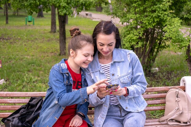 Mère et fille adolescente marchant dans un parc, assises sur un banc et regardant dans un smartphone, jeune femme caucasienne heureuse aux cheveux longs et adolescente surfant sur Internet à l'extérieur, famille de style de vie