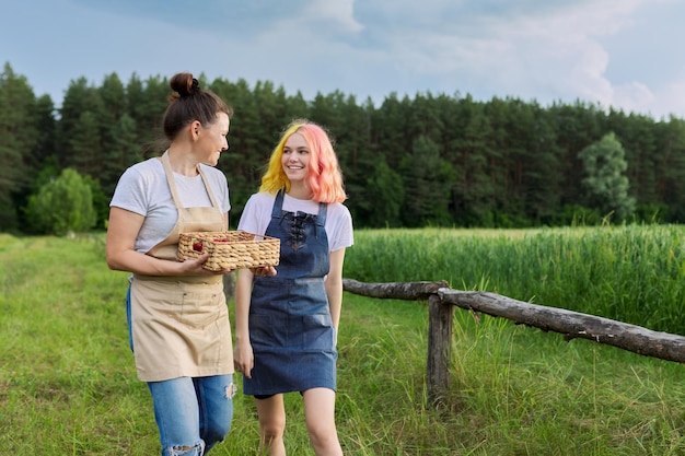 Mère et fille adolescente en tablier marchant avec panier de fraises fraîches, nature, ferme, beau fond de paysage. Alimentation biologique saine, travaux agricoles et loisirs