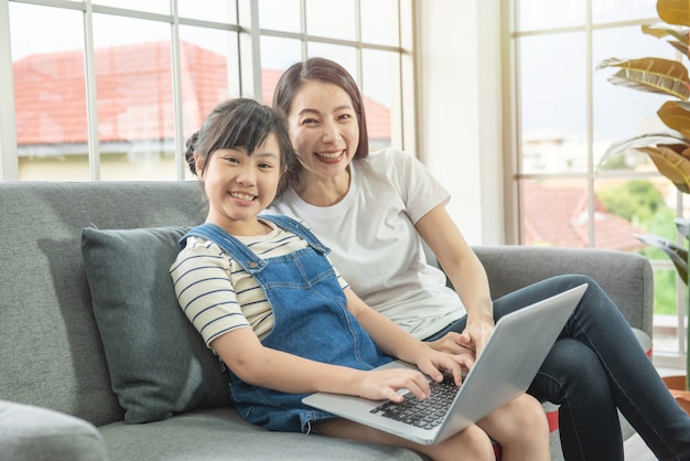 Mère et fille asiatique regarde la caméra avec le sourire s'asseoir sur le canapé au repos à l'aide d'un ordinateur portable en ligne