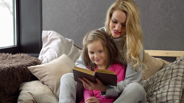 Mère et fille assises sur le lit dans une étreinte et lisant un livre.