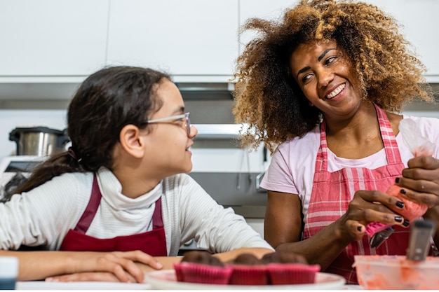 Mère et fille cuisinant des pâtisseries dans la cuisine femme adulte latine décorant des cupcakes avec un sac à poche rempli de crème sucrée rose véritable moment de liaison parent chilld