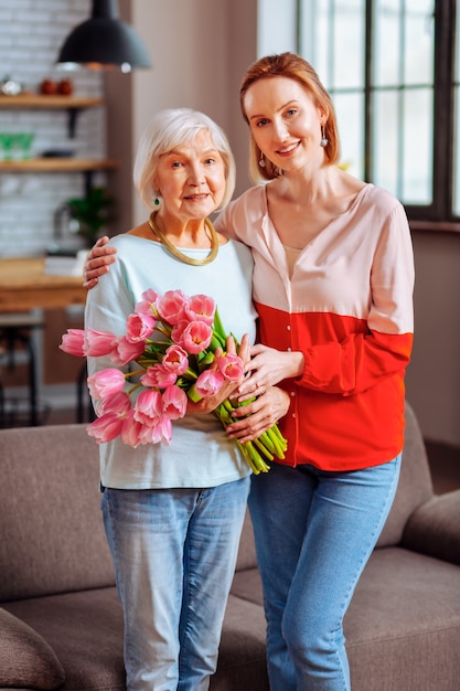 Mère Et Fille Avec Des Fleurs. Plan Complet D'une Dame Vieillissante Aux Cheveux Gris à La Retraite En Sweetshirt Bleu Et Jeans Tenant Des Tulipes Et Posant Avec Une Belle Dame Mature En Chemisier Rose