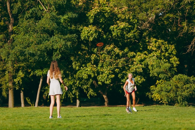 Mère et fille jetant un disque volant orange à un petit chien drôle, qui l'attrape sur l'herbe verte. Little Jack Russel Terrier animal jouant à l'extérieur dans le parc. Chien et femmes. Famille reposant en plein air