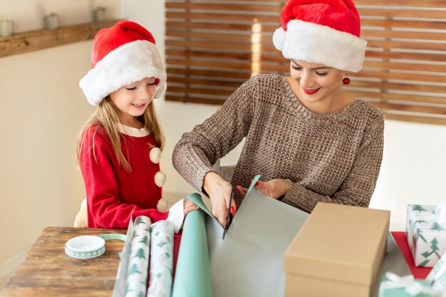 Photo mère et fille portant des chapeaux de père noël s'amusant à emballer des cadeaux de noël ensemble dans le salon