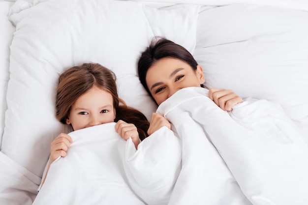 Mère et fille positives allongées dans leur lit tout en couvrant la moitié de leur visage avec une couverture blanche