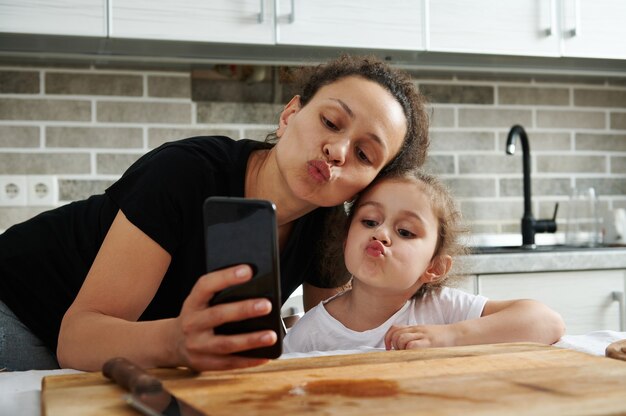 Mère et fille s'embrassant et regardant la caméra du téléphone portable tout en prenant un selfie dans la cuisine pendant la cuisson