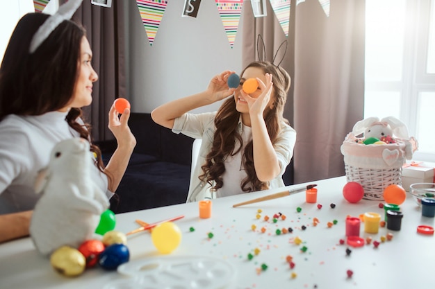 Mère Et Fille Se Préparent Pour Pâques. Fille Tenir Des œufs Colorés Devant Les Yeux. Mère Fait De Même. Ils S'amusent. Décoration Et Peinture Sur La Table.