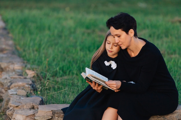 Mère et fille sont assises sur un banc de pierre et lisent un livre. Femme avec un enfant en robes noires.