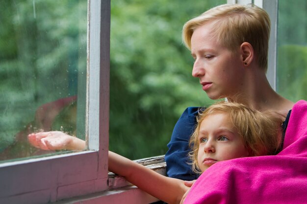 Mère et fille sont assises près de la fenêtre ouverte pendant la pluie, la mère tient sa fille dans une couverture sur elle-même