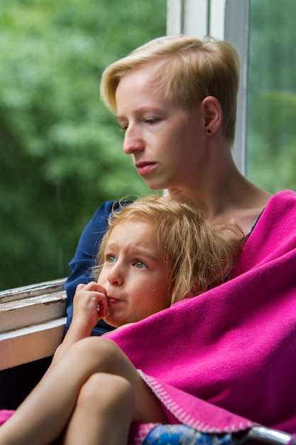 Mère et fille sont assises près de la fenêtre ouverte pendant la pluie, la mère tient sa fille dans une couverture sur elle-même