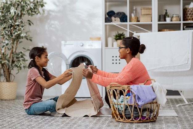 La mère et la fille sont assises sur le sol dans la buanderie de la maison, la fille aide la femme à faire les tâches ménagères, elle assemble des vêtements propres et lavés sortis d'un grand panier en osier