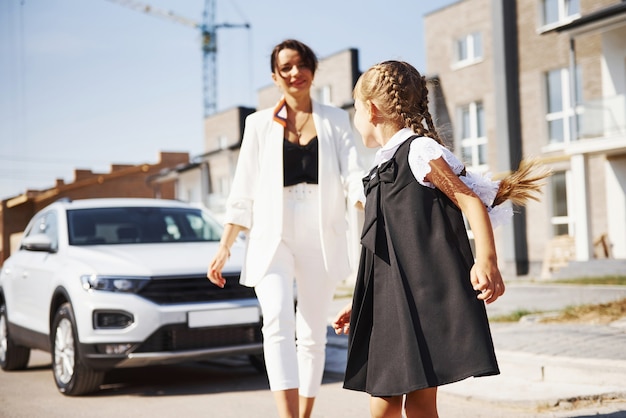 Mère avec fille en uniforme scolaire à l'extérieur près d'une voiture blanche.