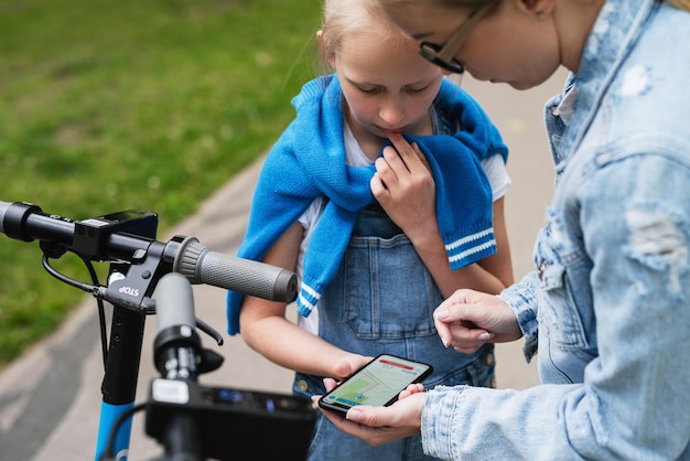 Mère et fille utilisant l'application sur smartphone pour partager des scooters électriques dans le parc de la ville