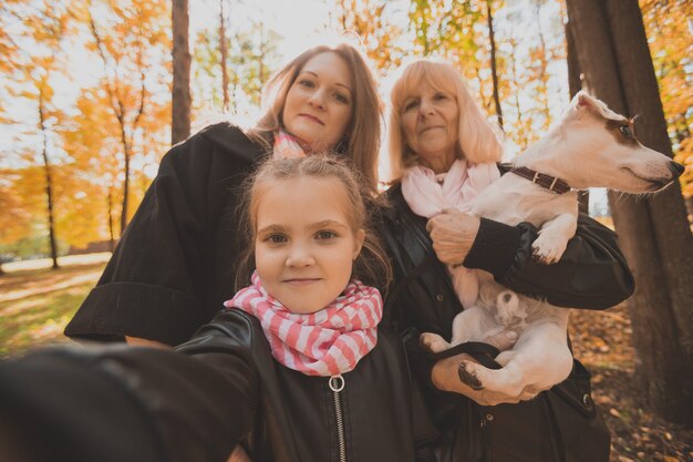 Mère, grand-mère et petite-fille avec chien jack russell terrier prenant selfie par smartphone à l'extérieur dans la nature d'automne. Concept de famille, d'animaux de compagnie et de génération