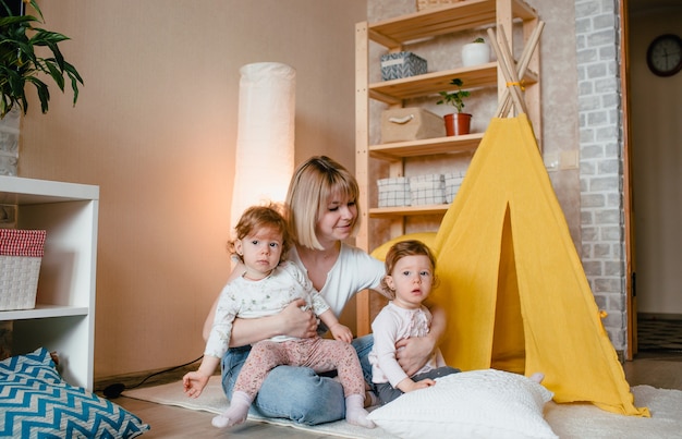 Une mère heureuse avec deux filles jumelles joue à la maison sur le sol assis près d'un tipi jaune. jeux conjoints de mères et de filles.