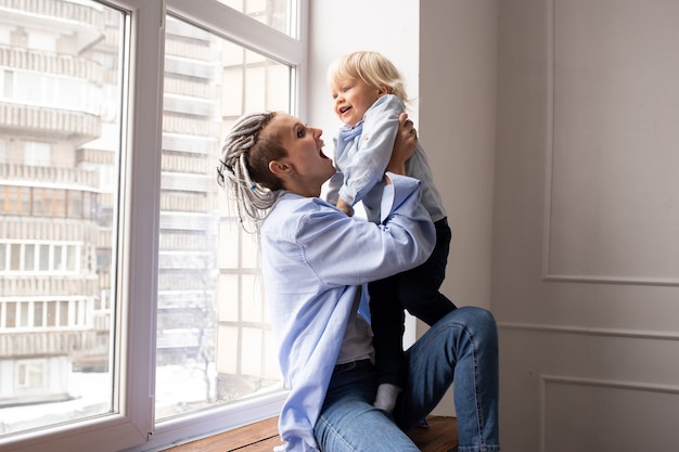 Mère hipster avec bébé garçon fils assis près de la fenêtre et jouant